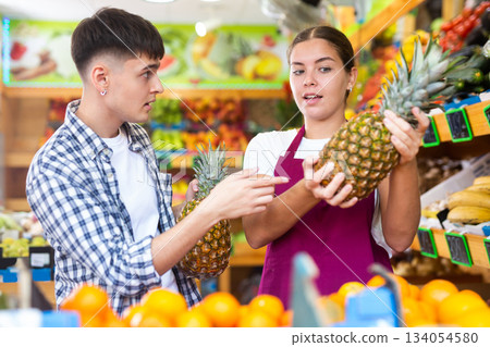 Friendly young saleswoman helps a guy buyer choose a pineapple Friendly young saleswoman helps a guy buyer choose a pineapple 134054580