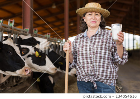 Portrait of an elderly farmer woman standing in a cowshed with a glass of milk 134054616