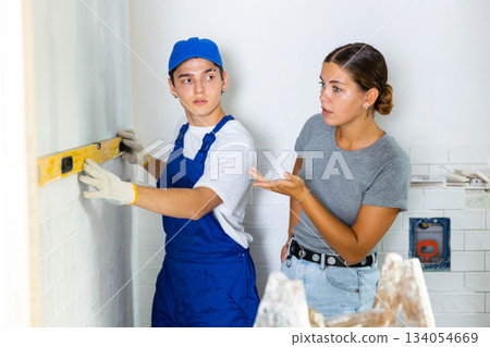 Young worker uses a spirit level on wall, consulting with a girl client 134054669