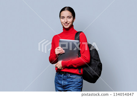 Portrait of beautiful female teenage student with backpack, laptop on gray background 134054847