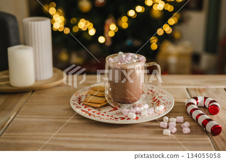 Holiday, Christmas background. A cup with hot chocolate and marshmallows on the table with Christmas tree on background Holiday, Christmas background. A cup with hot chocolate and marshmallows on the table with Christmas tree on background 134055058