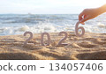 hand place the number 6 on sandy beach, transition from year 2025 to 2026 for new year sign on summer beach at sunset time, with ripple sea wave and beautiful nature at background, New Year celebrate 134057406