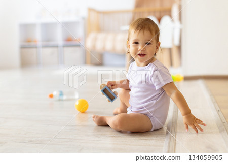 Smiling baby sitting on floor holding small toy in bright home interior 134059905