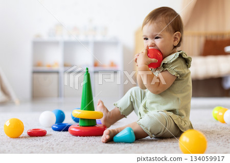 Infant baby biting colorful toy while sitting among scattered play items 134059917