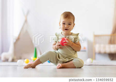 Baby sitting on floor holding red ball looking forward with curiosity Baby sitting on floor holding red ball looking forward with curiosity 134059932