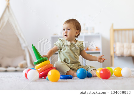 Cute infant baby girl sitting on soft carpet surrounded by colorful toys Cute infant baby girl sitting on soft carpet surrounded by colorful toys 134059933