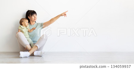 Mother sitting with baby on floor and pointing aside at copy space Mother sitting with baby on floor and pointing aside at copy space 134059937