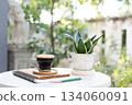Black coffee and potted plant and stack of notebooks on white table with garden view 134060091