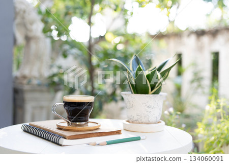 Black coffee and potted plant and stack of notebooks on white table with garden view Black coffee and potted plant and stack of notebooks on white table with garden view 134060091