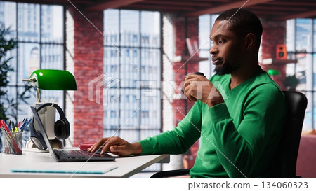 African american adult typing on a computer in his loft apartment, engaging in online freelancing work and multitasking efficiently. Flexibility of remote work in a cozy home office. Camera A. African american adult typing on a computer in his loft apartment, engaging in online freelancing work and multitasking efficiently. Flexibility of remote work in a cozy home office. Camera A. 134060323