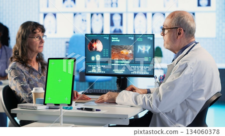 Senior medic consulting ill woman in cabinet with isolated mockup screen, checking the hospital records to do another x ray scan interpretation. Old patient discuss with her physician. Camera B. 134060378