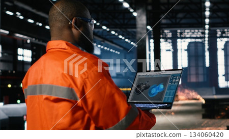 Technician in a manufacturing facility uses automation software on laptop to manage a turbine system surrounded by heavy equipment, steel machinery and fabrication tools. 134060420