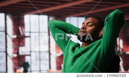 Man at home deals with back pain and stretching joints on chair, being uncomfortable due to incorrect posture. Black guy feeling overworked and experiencing muscle cramps at desk. Camera A. Man at home deals with back pain and stretching joints on chair, being uncomfortable due to incorrect posture. Black guy feeling overworked and experiencing muscle cramps at desk. Camera A. 134060588