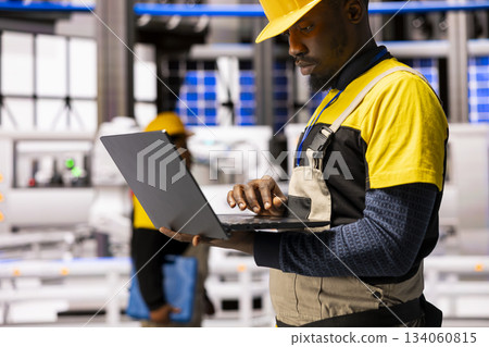 Black male engineer enhances assembly line efficiency by managing automation software on laptop. Industrial technician assesses robot arm manufacture and industry 4.0 systems in automated factory. Black male engineer enhances assembly line efficiency by managing automation software on laptop. Industrial technician assesses robot arm manufacture and industry 4.0 systems in automated factory. 134060815