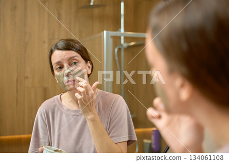Woman applying clay face mask and looking at reflection in bathroom mirror 134061108