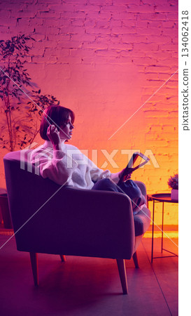 Woman sitting with tablet in cozy neon environment holding pen and thinking. 134062418