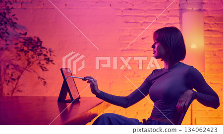 Woman pointing at tablet screen with stylus in neon workspace. Woman pointing at tablet screen with stylus in neon workspace. 134062423