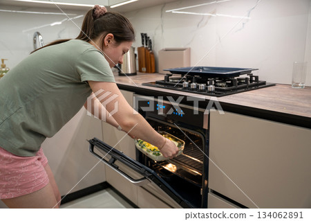 Young caucasian brunette woman places a casserole with broccoli, red fish and cheese in the oven. Young caucasian brunette woman places a casserole with broccoli, red fish and cheese in the oven. 134062891