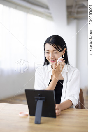 一位女士坐在窗邊的桌子上，對著鏡子用粉撲化妝。 134063001