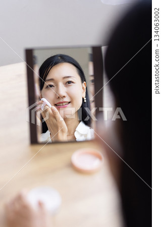 一位女士坐在窗邊的桌子上，對著鏡子用粉撲化妝。 134063002