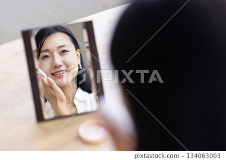 一位女士坐在窗邊的桌子上，對著鏡子用粉撲化妝。 134063003