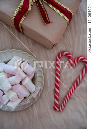 Flat-lay top view showing half of a festive mug filled with marshmallows and half of a wrapped gift box decorated with red-and-white heart-shaped candy canes. Cozy holiday composition perfect for Flat-lay top view showing half of a festive mug filled with marshmallows and half of a wrapped gift box decorated with red-and-white heart-shaped candy canes. Cozy holiday composition perfect for 134065694