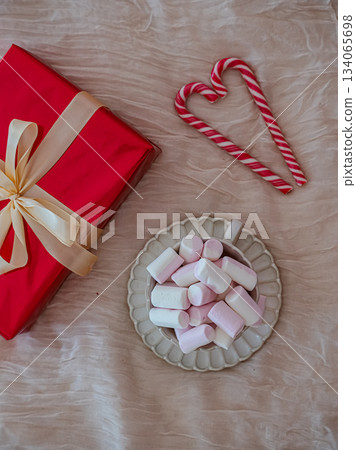 Flat-lay top view of a festive holiday mug filled with marshmallows placed next to a wrapped gift box decorated with red-and-white heart-shaped candy canes. Cozy Christmas composition suitable for 134065698