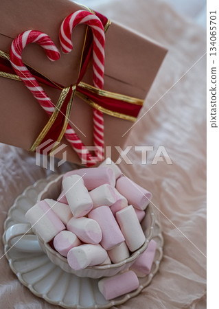 Photo showing a festive mug filled with marshmallows and a wrapped gift box decorated with red-and-white heart-shaped candy canes tied with a ribbon. Cozy holiday composition ideal for greeting cards Photo showing a festive mug filled with marshmallows and a wrapped gift box decorated with red-and-white heart-shaped candy canes tied with a ribbon. Cozy holiday composition ideal for greeting cards 134065701
