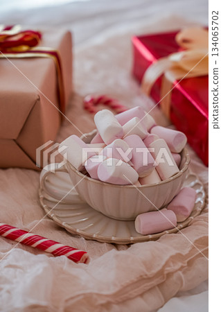 Close-up side view of a festive mug filled with marshmallows, with wrapped Christmas gift boxes softly blurred in the background. Cozy holiday composition ideal for seasonal marketing, greeting cards Close-up side view of a festive mug filled with marshmallows, with wrapped Christmas gift boxes softly blurred in the background. Cozy holiday composition ideal for seasonal marketing, greeting cards 134065702