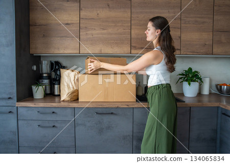 Young woman unpacking a box with pre-portioned ingredients for cooking on the kitchen counter. Concept of meal kit delivery service and easy home cooking with fresh food. 134065834