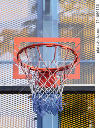 An orange metal basketball rim and net are mounted on a fiberglass backboard, prominently framed by a steel grid fence. An orange metal basketball rim and net are mounted on a fiberglass backboard, prominently framed by a steel grid fence. 134066186