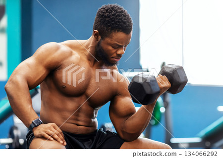 Handsome african american muscular shirtless man bodybuilder exercising with barbell at gym, sitting on bench and looking at biceps, copy space. Gym, male gym, fitness studio advertisement 134066292