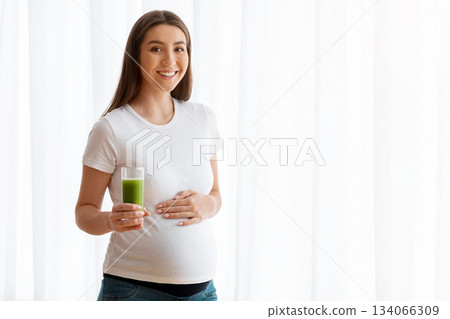 Portrait Of Smiling Pregnant Woman With Glass Of Green Vegetable Smoothie Standing Near Window At Home, Young Lady Enjoying Organic Vitamin Juice Drink For Healthy Pregnancy, Copy Space Portrait Of Smiling Pregnant Woman With Glass Of Green Vegetable Smoothie Standing Near Window At Home, Young Lady Enjoying Organic Vitamin Juice Drink For Healthy Pregnancy, Copy Space 134066309