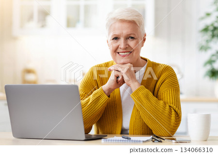Happy elderly lady waiting for webinar, sitting in kitchen, using laptop and taking notes, drinking coffee. Cheerful aged woman grandmother having educational class on Internet, copy space Happy elderly lady waiting for webinar, sitting in kitchen, using laptop and taking notes, drinking coffee. Cheerful aged woman grandmother having educational class on Internet, copy space 134066310