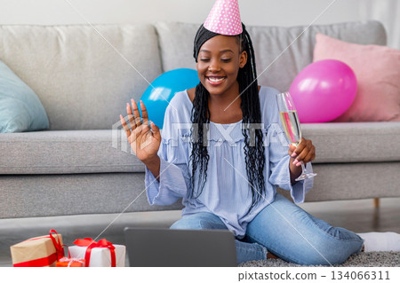 Happy african american young lady with glass of champagne and festive hat having online party from home, using laptop, sitting on floor next to giftboxes and colorful balloons, copy space Happy african american young lady with glass of champagne and festive hat having online party from home, using laptop, sitting on floor next to giftboxes and colorful balloons, copy space 134066311