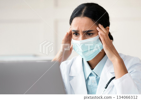 Exhausted arab female doctor in protective face mask working in clinic, sitting at workdesk in front of laptop, touching her head, need for break, copy space. Brunette woman physician having headache Exhausted arab female doctor in protective face mask working in clinic, sitting at workdesk in front of laptop, touching her head, need for break, copy space. Brunette woman physician having headache 134066312
