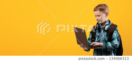 Technologies in school education. Smart schoolboy with backpack and headset holding laptop over yellow studio background, copy space. Cute kid using laptop for studying, doing homework, websurfing 134066313