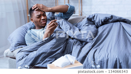 Sick African Man Having Fever Touching Forehead Holding Thermometer Lying On Couch At Home. Ill Black Guy Measuring Temperature. Influenza Symptoms Concept. Selective Focus Sick African Man Having Fever Touching Forehead Holding Thermometer Lying On Couch At Home. Ill Black Guy Measuring Temperature. Influenza Symptoms Concept. Selective Focus 134066343