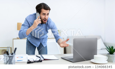 Angry Businessman Talking On Phone Looking At Laptop, Having Computer Problem Standing In Office. Communication Issue, Worlplace Problems And Conflicts Concept. Panorama, Selective Focus Angry Businessman Talking On Phone Looking At Laptop, Having Computer Problem Standing In Office. Communication Issue, Worlplace Problems And Conflicts Concept. Panorama, Selective Focus 134066345