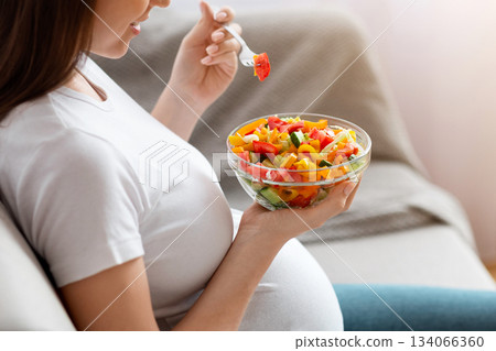 Young Pregnant Woman Eating Fresh Vegetable Salad For Lunch, Sitting On Sofa At Home, Enjoying Healthy Organic Food And Rich Vitamin Nutrition During Pregnancy, Side View Shot With Free Space Young Pregnant Woman Eating Fresh Vegetable Salad For Lunch, Sitting On Sofa At Home, Enjoying Healthy Organic Food And Rich Vitamin Nutrition During Pregnancy, Side View Shot With Free Space 134066360