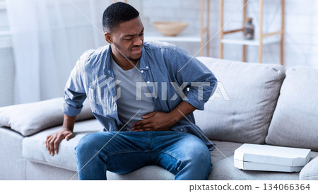 African American Guy Having Stomachache After Eating Pizza Touching Aching Stomach Suffering From Pain Sitting On Sofa At Home. Food Poisoning, Gastritis, Abdominal Inflammation And Appendicitis. African American Guy Having Stomachache After Eating Pizza Touching Aching Stomach Suffering From Pain Sitting On Sofa At Home. Food Poisoning, Gastritis, Abdominal Inflammation And Appendicitis. 134066364
