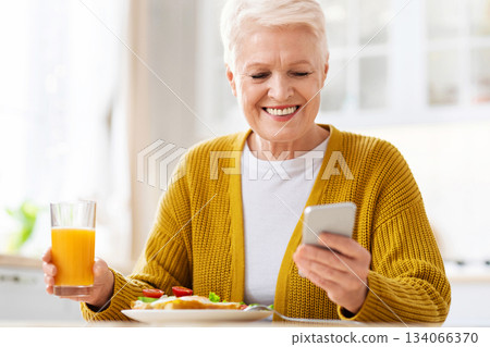 Cheerful elderly woman grandmother using mobile phone while having lunch, eating sandwich and drinking juice, sitting alone at kitchen, empty space. Senior woman with smartphone at home, closeup 134066370