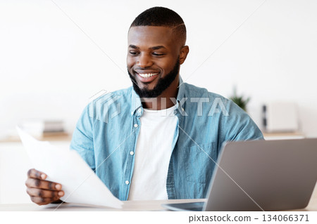 Millennial Black Freelancer Man Working With Papers At Desk In Home Office. Smiling African American Guy Sitting At Table With Computer Reading Documents And Checking Financial Reports, Free Space Millennial Black Freelancer Man Working With Papers At Desk In Home Office. Smiling African American Guy Sitting At Table With Computer Reading Documents And Checking Financial Reports, Free Space 134066371