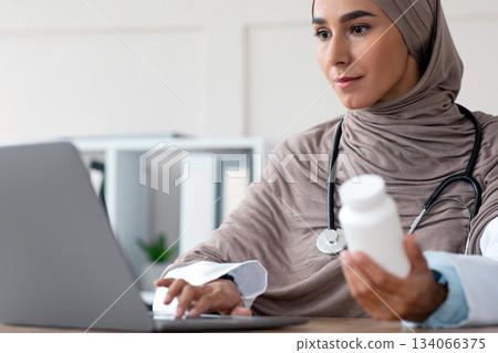 Closeup of female doctor young woman in hijab holding jar with medicine supplements or vitamins and using laptop, muslim lady physician working online from office in clinic, copy space 134066375