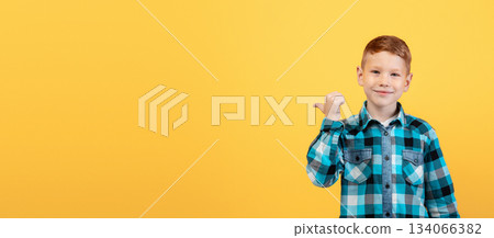 Smiling red-haired caucasian boy aiming at free space behind his back, empty space for text or advertisement, panorama. Cheerful ginger kid pointing with thumb at copy space over yellow background Smiling red-haired caucasian boy aiming at free space behind his back, empty space for text or advertisement, panorama. Cheerful ginger kid pointing with thumb at copy space over yellow background 134066382