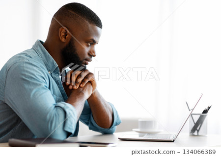 Thoughtful Black Writer Working With Laptop At Home Office. African American Freelancer Man Sitting At Desk And Looking At Computer Screen With Pensive Face Expression, Side View With Copy Space Thoughtful Black Writer Working With Laptop At Home Office. African American Freelancer Man Sitting At Desk And Looking At Computer Screen With Pensive Face Expression, Side View With Copy Space 134066389