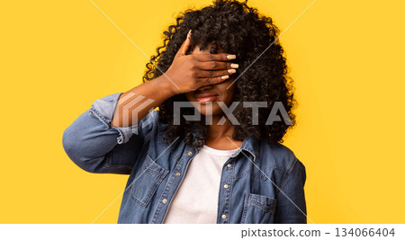 Curly black woman covering eyes with one hand, feeling scared or anxious, wondering or blindly waiting for a surprise against yellow studio background, panorama with copy space 134066404