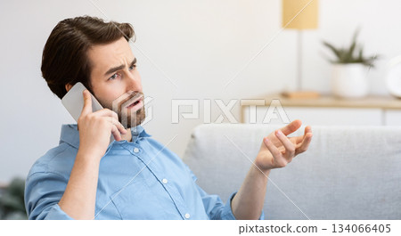 Discontented Guy Frowning Talking On Cellphone Sitting With Laptop, Having Computer Or Internet Connection Problem At Home. Displeased Customer Calling Hotline. Remote Work Issue. Selective Focus 134066405