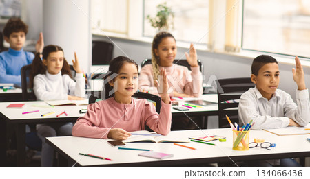 First Day Of School. Multiethnic group of diverse little classmates sitting at tables in modern classroom, raising hands up for an answer, discussing new theme with teacher, participation concept First Day Of School. Multiethnic group of diverse little classmates sitting at tables in modern classroom, raising hands up for an answer, discussing new theme with teacher, participation concept 134066436