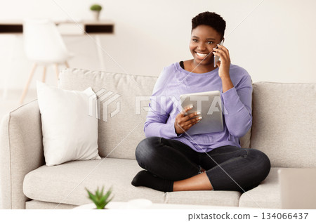 Always In Touch. Happy young plus size african american woman talking on mobile phone, sitting on the couch at home and holding digital tablet. Communication And Technology Concept. Always In Touch. Happy young plus size african american woman talking on mobile phone, sitting on the couch at home and holding digital tablet. Communication And Technology Concept. 134066437